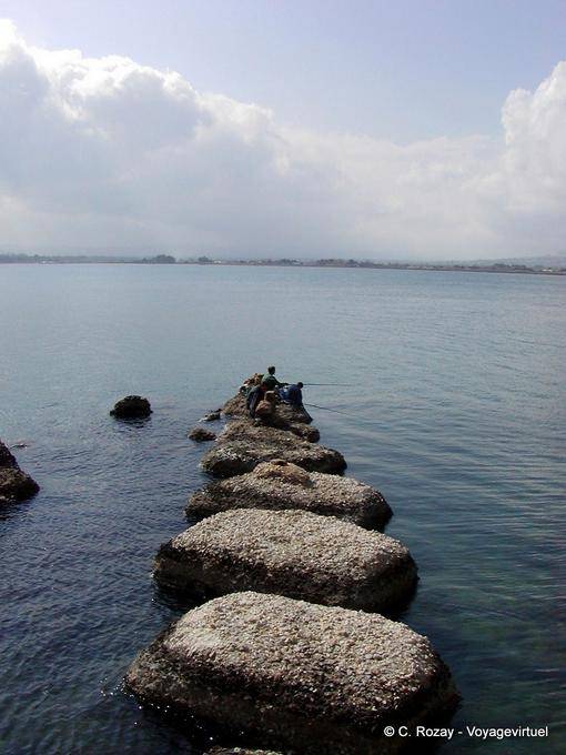 Pescadores en el muelle de piedras volcánicas negras, Siracusa, Sicilia - Italia